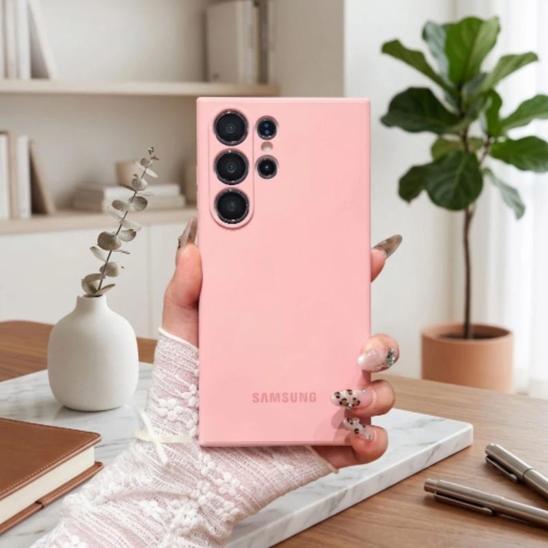 May include: A light pink smartphone case with a smooth matte finish, featuring a rectangular cutout for the camera array on the upper left side and the word SAMSUNG printed in a matching pink tone near the bottom center of the back panel.