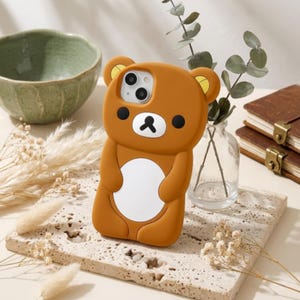 May include: A brown silicone phone case shaped like a bear with rounded ears, a white circular belly patch, and a face featuring black eyes and a black nose. The case has a cutout for a dual-lens camera system on the upper left side.