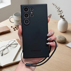 May include: A matte black smartphone case featuring a rectangular design with a multi-lens camera array at the top left corner and the Samsung logo embossed at the bottom. The case includes a black wrist strap attached to the side for carrying.