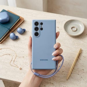 May include: A light blue Samsung smartphone case with a matching wrist strap attached to the bottom. The case features a rectangular design with a cutout for the rear camera array and the Samsung logo printed in the center of the back panel.