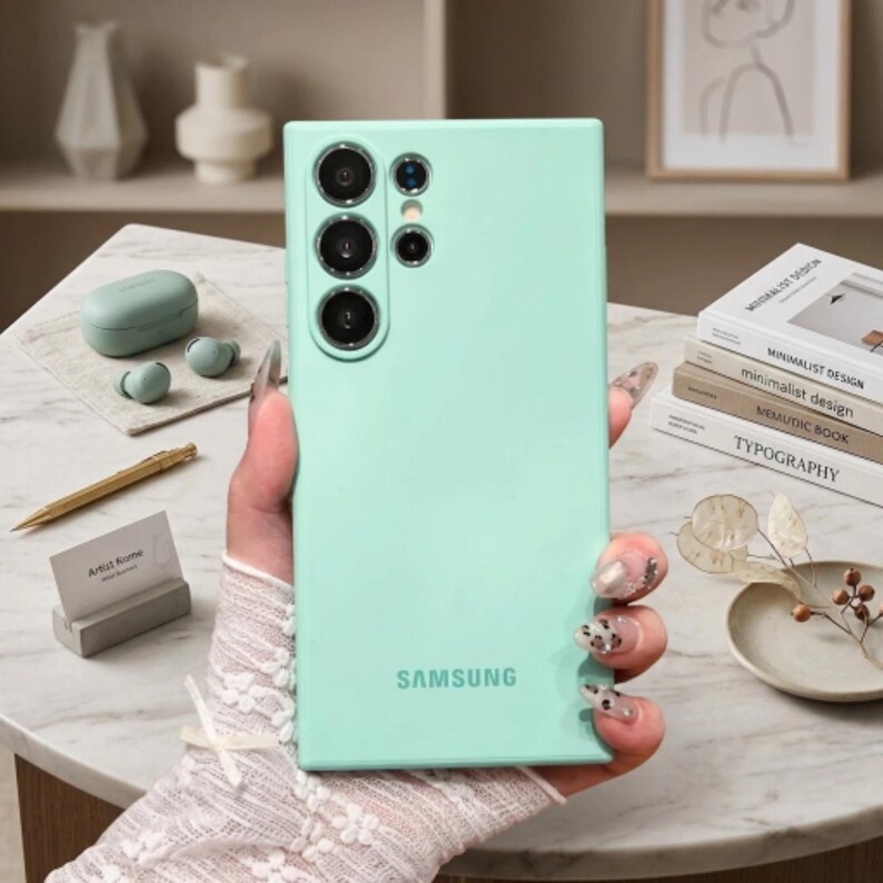 May include: A mint green smartphone case featuring a rectangular shape with rounded corners and a vertical camera array on the back. The Samsung logo is printed in a subtle font at the bottom center of the smooth, matte-textured protective cover.