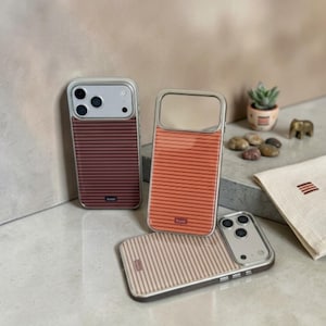 May include: Three rectangular smartphone cases featuring horizontal pinstripes in shades of dark red, orange, and beige. Each case has a clear camera cutout area and a small rectangular logo tag at the bottom. The cases are arranged in a group.