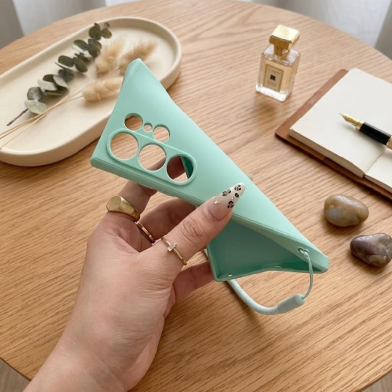 May include: A mint green smartphone case featuring a series of circular cutouts for the camera lenses on the back panel. The case includes a matching mint green wrist strap attached to the bottom corner, providing a secure way to carry the device.