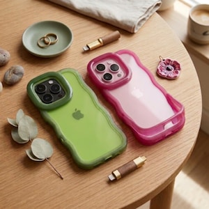 Puede incluir: Dos fundas para smartphone con bordes ondulados, una en verde lima translúcido y otra en magenta translúcido, cada una con un recorte elevado para la cámara. Las fundas se muestran junto a dos cables de carga de madera, un pequeño plato verde con dos anillos dorados y un dije floral.