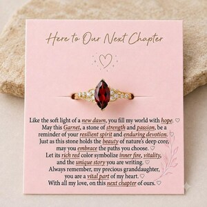 Könnte beinhalten: Ein goldener Ring mit einem tiefroten Granatstein im Marquise-Schliff, flankiert von kleinen klaren Schmucksteinen, befestigt auf einer hellrosa Karte mit dem Text Here to Our Next Chapter und einer Nachricht an eine Enkelin.