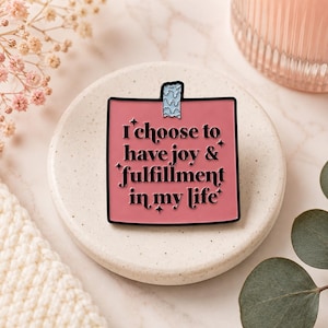 May include: A square pink enamel pin shaped like a sticky note with a small piece of blue patterned tape at the top. The pin features black text that reads I choose to have joy & fulfillment in my life, accented by small black star symbols.