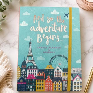 May include: A teal travel planner and journal with a gold elastic band. The cover features a cityscape illustration with the Eiffel Tower and a Ferris wheel, topped with the rose gold foil text: And so the adventure Begins... TRAVEL PLANNER & JOURNAL.