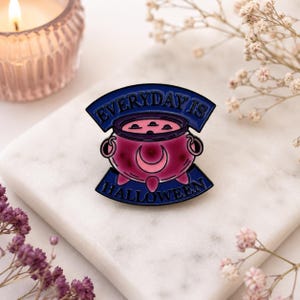 May include: A metal enamel pin featuring a pink cauldron with a crescent moon design, surrounded by a dark blue banner with the text EVERYDAY IS HALLOWEEN in black lettering. The pin has a glossy finish and a distinct, rounded shape.