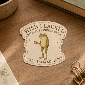May include: A die-cut sticker with a cream background featuring a green frog holding a small orange mug. The text reads WISH I LACKED CRITICAL THINKING SKILLS Y'ALL SEEM SO HAPPY in brown lettering, accented by four small brown star symbols.