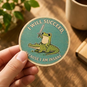 Puede incluir: Una pegatina circular con fondo verde azulado que muestra una rana verde montada sobre un caimán verde mientras sostiene una espada plateada. El texto I WILL SUCCEED, BECAUSE I AM INSANE está impreso en color crema en los bordes.