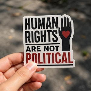 Peut inclure: Un autocollant rectangulaire à fond blanc avec le texte noir HUMAN RIGHTS, le texte noir ARE NOT et le texte rouge POLITICAL. À droite, une silhouette noire de main ouverte contient un petit cœur rouge au centre de la paume.