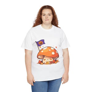 May include: A white t-shirt featuring a graphic of a cartoon mushroom with an orange cap and white spots. The mushroom is surrounded by autumn leaves and holds a purple flag with a jack-o'-lantern face. The mushroom wears small yellow boots.