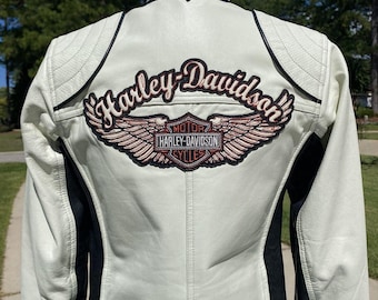 Könnte beinhalten: Eine weiß-schwarze Motorradjacke aus Leder mit einem großen gestickten Harley-Davidson-Logo auf dem Rücken. Das Design zeigt den Harley-Davidson-Schriftzug über einem geflügelten Emblem mit den Worten Motor Harley-Davidson Cycles.