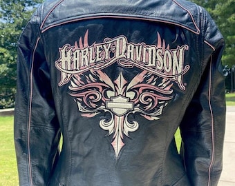 Peut inclure: Une veste en cuir noir avec un grand logo Harley-Davidson brodé dans le dos. Le motif comprend des ailes stylisées roses et blanches ainsi qu'un emblème central en forme de bouclier. La veste présente un liseré rose le long des coutures.