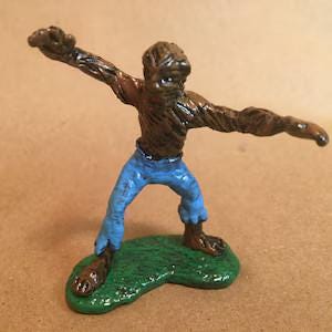 May include: A small, hand-painted figurine of a brown, textured creature with a humanoid shape, wearing bright blue pants. The figure is posed with arms outstretched in a dynamic stance and stands on a textured, oval-shaped green base.