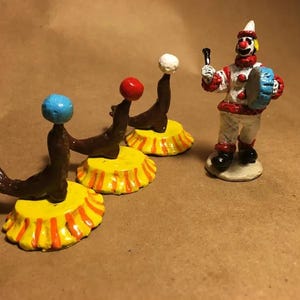 May include: Two brown seal figurines balancing colorful balls on their noses, each standing on a yellow and orange striped circular base, accompanied by a clown figurine wearing a white and red outfit while holding a blue drum and a black mallet.