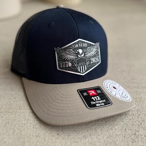 May include: A navy blue and tan baseball cap with a black mesh back. The front features a hexagonal patch with an eagle, the text 250 YEARS, 1776, 2026, and MADE IN AMERICA. A black sticker on the brim displays the text 112 The Original MD-LG.