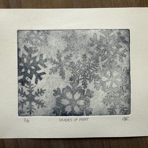 May include: A rectangular print on off-white paper featuring dark blue snowflake patterns of varying sizes and shapes. The print is titled SHADES OF FROST, marked 2/8, and signed with the initials AR in the bottom margin of the paper.