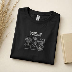 May include: A black t-shirt featuring a white graphic with the text "THINGS I DO IN MY SPARE TIME" above six icons representing pottery activities: making pottery, buying clay, watching pottery videos, talking, thinking, and dreaming about pottery.
