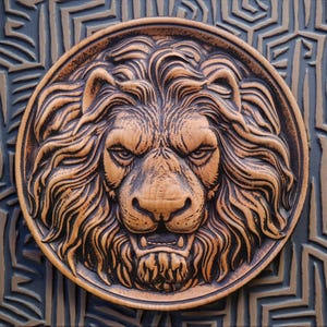 Könnte beinhalten: Eine kreisförmige Wandplatte mit einem Hochrelief eines Löwenkopfes mit voller Mähne. Der Löwe hat einen konzentrierten Ausdruck mit entblößten Zähnen. Das Stück ist in einem hellen Beigeton mit dunkelbraunen Schattierungen in den Vertiefungen.