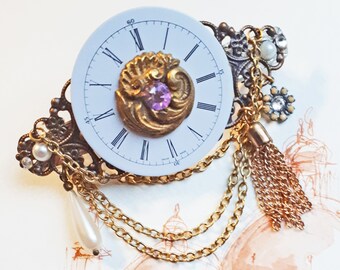 May include: A vintage brooch with a white clock face, black Roman numerals, and gold-coloured filigree. It features a central purple gemstone, gold chains, and a tassel. The brooch is adorned with pearls and other decorative elements.