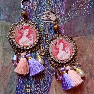 Peut inclure: Boucles d'oreilles pendantes ornées d'un portrait de femme rousse. Les boucles d'oreilles ont un cadre en filigrane de couleur bronze avec des strass transparents et sont ornées de pompons pêche et violets, de perles transparentes et nacrées.