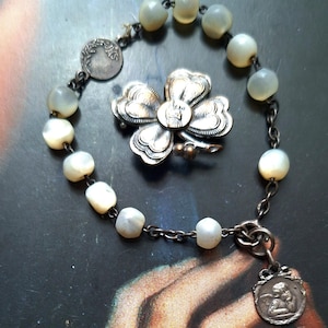 May include: An antique bracelet featuring off-white, pearlescent beads and a silver-toned chain. A silver flower brooch and a small angel pendant are also visible. The jewellery is displayed on a dark, textured surface.