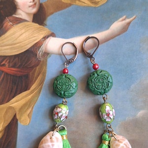 Op de afbeelding: Dangel oorbellen met groene gebeeldhouwde stenen kralen, bloemen emaille kralen, kleine groene kwastjes en schelpen. De oorbellen hebben koperkleurige oorhaken en rode kralen.