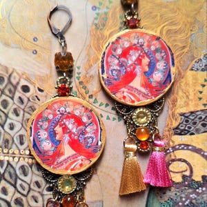 Peut inclure: Boucles d'oreilles de style Art nouveau avec des pendentifs ronds représentant le profil d'une femme en rouge, bleu et or. Les boucles d'oreilles ont des ornements métalliques décoratifs, des pierres précieuses colorées et des pompons dorés et roses. Le fond a un motif or et vert.