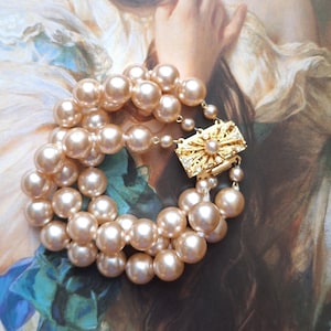 Könnte beinhalten: Ein Armband im Vintage-Stil mit mehreren Strängen großer, pfirsichfarbener Kunstperlen. Das Armband wird mit einem goldfarbenen Verschluss gesichert, der mit einer zentralen Perle und klaren Kristallen verziert ist. Der Hintergrund zeigt ein Gemälde mit sanften Farben.