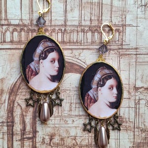 Peut inclure: Paire de boucles d'oreilles dorées avec des pendentifs ovales représentant un portrait de femme. Chaque boucle d'oreille a un fermoir à levier doré, une perle de cristal grise et un groupe d'étoiles en bronze, une perle en forme de goutte et un charme de plume. Mesure environ 7 cm de long.