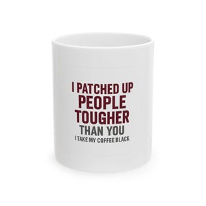 May include: A white ceramic mug featuring dark red and gray text that reads: I PATCHED UP PEOPLE TOUGHER THAN YOU I TAKE MY COFFEE BLACK. The mug has a cylindrical shape with a smooth finish and is presented against a plain, neutral background.