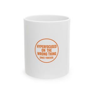 May include: A white ceramic mug featuring an orange circular graphic on the front. Inside the circle, the text reads in orange capital letters: HYPERFOCUSED ON THE WRONG THING SINCE FOREVER. The mug has a smooth, cylindrical body and a simple handle.
