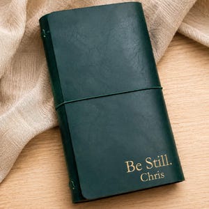 May include: A dark green leather journal with a smooth, matte finish. The cover features the gold-embossed text "Be Still. Chris" in the bottom right corner. A thin, matching green elastic band wraps horizontally around the centre of the notebook.