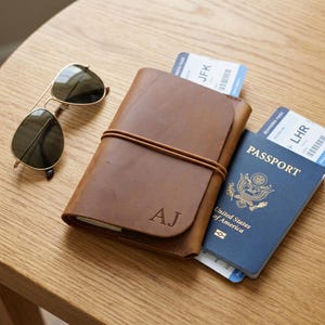 Può includere: Un porta passaporto in pelle color cuoio con le iniziali AJ impresse sul davanti, fissato da un sottile elastico. Accanto si trovano occhiali da sole aviatore scuri e un passaporto statunitense blu con due carte d'imbarco per JFK e LHR.
