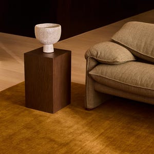 May include: A speckled off-white ceramic bowl with a pedestal base sits atop a dark brown wooden rectangular side table. The bowl has a rounded shape and a textured surface, positioned next to a textured tan sofa on a golden brown rug.