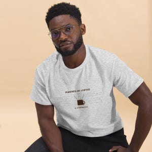 Peut inclure: Un t-shirt gris clair à manches courtes avec un motif représentant une tasse de café marron dont la vapeur forme un circuit imprimé. Le texte au-dessus de la tasse indique POWERED BY COFFEE et en dessous & SCHEMATICS en noir.