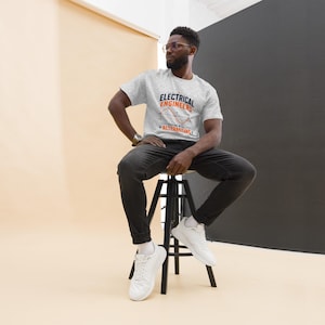 Peut inclure: Un t-shirt gris clair à manches courtes avec le texte "ELECTRICAL ENGINEERS" en bleu foncé et "ALTERNATING" en orange, accompagné d'un graphique d'onde sinusoïdale. Le t-shirt est porté avec un jean gris foncé et des baskets blanches.