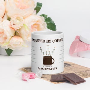 Peut inclure: Un mug en céramique blanc avec le texte POWERED BY COFFEE & SCHEMATICS en lettres noires. Le motif comprend un graphique d'une tasse à café marron avec un schéma de circuit électronique qui en sort, ainsi que le petit texte LAB MODE ON.