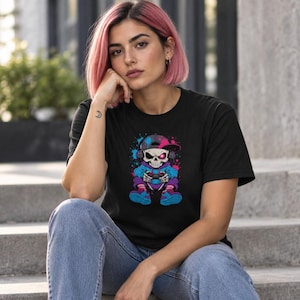 Op de afbeelding: Een zwart t-shirt met korte mouwen en een grafische print van een cartoon-skelet met een blauwe hoodie, een roze en zwarte honkbalpet en een koptelefoon, terwijl het een gamecontroller vasthoudt, geaccentueerd door roze en blauwe verfspatten.