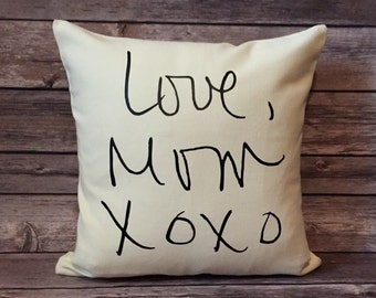 May include: Cream-colored square pillow with the handwritten text "Love, Mom xoxo" in black. The pillow is placed on a wooden surface with a rustic, weathered appearance. The pillow is a decorative item.
