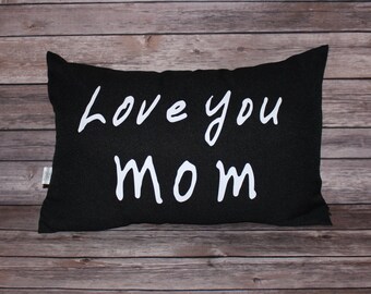 May include: Black rectangular pillow with the words "Love you Mom" in white script. The pillow is made of a textured fabric and is displayed on a wooden surface.