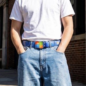 Könnte beinhalten: Ein weißes T-Shirt mit Rundhalsausschnitt, kombiniert mit einer hellblauen Jeans und einem blauen Gürtel mit quadratischer Schnalle, die ein buntes, viergeteiltes Fenster-Logo zeigt. Der Gürtel steckt in den Schlaufen der klassisch geschnittenen Jeans.