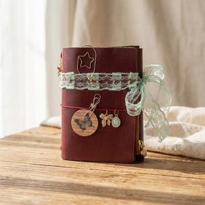 May include: A dark red leather journal featuring a mint green lace ribbon tied around the cover. It is decorated with a star-shaped paperclip, a circular tag with a butterfly illustration, and two small metal charms shaped like a bow and an oval.