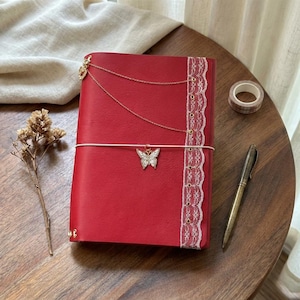 May include: A red journal featuring a white lace trim along the right edge, a thin white elastic band closure, and a silver butterfly charm. The cover is adorned with two delicate gold-toned chains attached to the top left corner of the notebook.