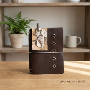 May include: A dark brown leather journal featuring a vertical row of five silver star-shaped metal eyelets on the front cover. A silver star-shaped metal clip holds a piece of vintage-style paper with printed text and a feather illustration.