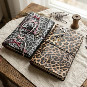Può includere: Due quaderni rettangolari con copertine leopardate. Il quaderno a sinistra presenta una stampa leopardata grigia, lacci in nastro rosa e una clip metallica a forma di stella. Quello a destra ha una stampa leopardata marrone e ciondoli.