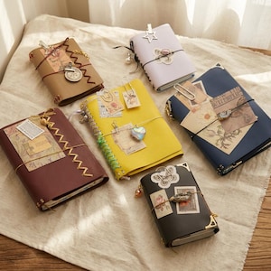 May include: Six rectangular journals in various colors including tan, lavender, yellow, dark blue, maroon, and black. Each journal is decorated with paper ephemera, metal clips, charms, and elastic bands. The covers feature textured surfaces and metal corner accents.