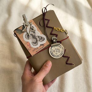 May include: A tan notebook featuring a dark maroon ribbon laced through metal eyelets. It is decorated with a silver star clip, a butterfly sticker, a gold chain, and a circular metal tag stamped with text including ISTANBUL and 02 19 03.