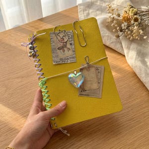 May include: A bright yellow notebook with a textured cover, featuring a lavender and lime green ribbon lacing along the side. It is decorated with a silver star paperclip, a plain silver paperclip, and a dangling iridescent heart charm.